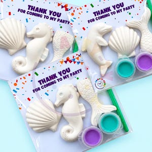 May include: Three craft kits in clear packaging, each featuring white sea-themed plaster figures like seahorses, shells, and mermaid tails. Each kit includes two small paint pots in teal and purple, a green paintbrush, and a card reading THANK YOU FOR COMING TO MY PARTY.