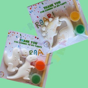 Peut inclure: Deux kits de peinture de dinosaures. Chaque kit comprend deux figurines de dinosaures blancs, deux pots de peinture (orange et vert, jaune et vert) et un pinceau. L'emballage indique "THANK YOU FOR COMING TO MY PARTY" et "ROAR!"