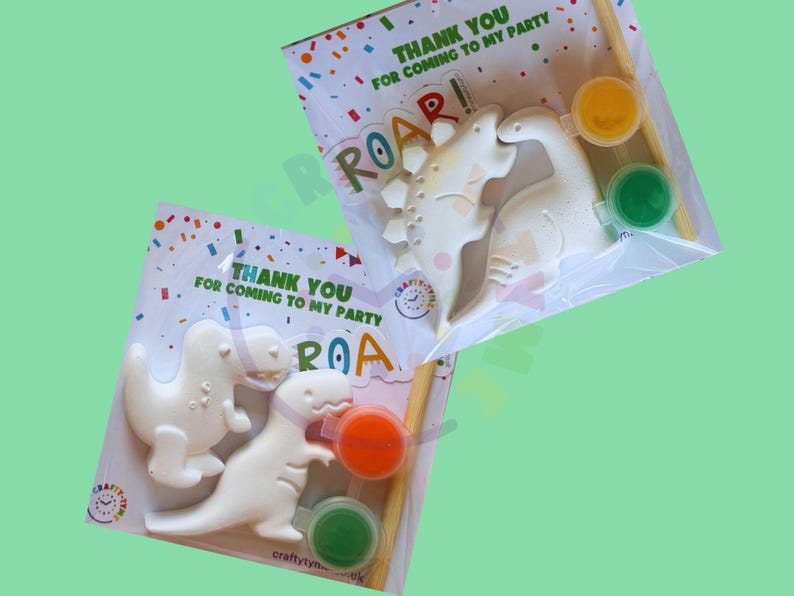 Peut inclure: Deux kits de peinture de dinosaures. Chaque kit comprend deux figurines de dinosaures blancs, deux pots de peinture (orange et vert, jaune et vert) et un pinceau. L'emballage indique "THANK YOU FOR COMING TO MY PARTY" et "ROAR!"