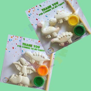 Peut inclure: Kits de bricolage &agrave; peindre soi-m&ecirc;me sur le th&egrave;me des dinosaures. Chaque kit comprend plusieurs figurines de dinosaures, des pots de peinture jaune, orange et vert, et un pinceau. L'emballage contient le texte "THANK YOU FOR COMING TO MY PARTY" avec des confettis color&eacute;s.