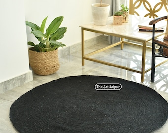 Puede incluir: Una alfombra redonda de yute negro sobre un suelo embaldosado. En el fondo, una silla decorativa con un cojín estampado, una mesa con tapa de mármol y plantas en macetas. La alfombra es un elemento clave en un salón.