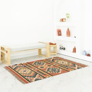 May include: A rectangular rug with a red border and geometric patterns in grey, black, and tan. It lies in front of a wooden bench with a grey cushion. A small wooden side table is positioned beside the bench. The rug's design is intricate.