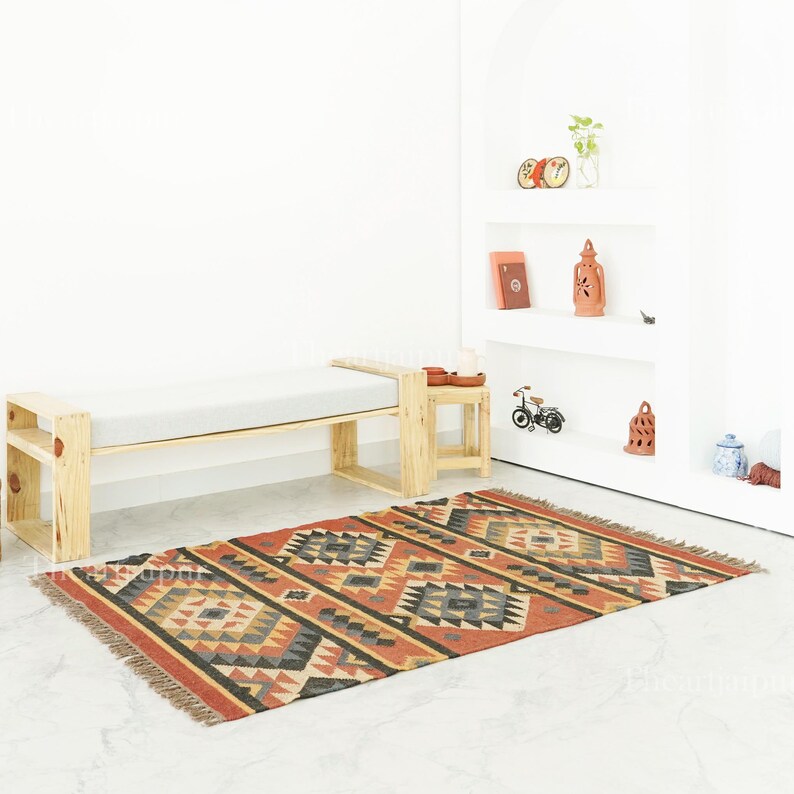 May include: A rectangular rug with a red border and geometric patterns in grey, black, and tan. It lies in front of a wooden bench with a grey cushion. A small wooden side table is positioned beside the bench. The rug's design is intricate.