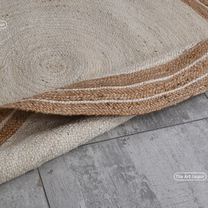 K&ouml;nnte beinhalten: Ein runder Jute-Teppich in naturbeige mit einem braunen und wei&szlig;en Streifenrand. Der Teppich ist zur H&auml;lfte gefaltet und der Rand ist sichtbar. Der Text "The Art Jaipur" ist auf dem Teppich sichtbar.