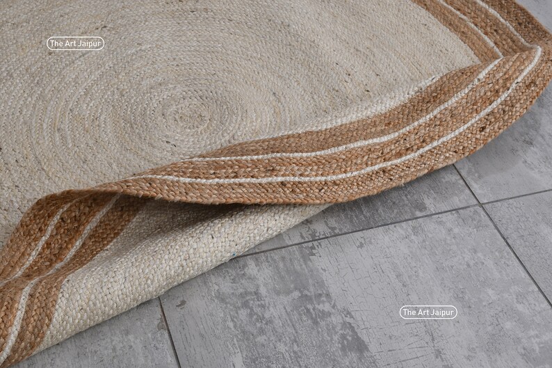 K&ouml;nnte beinhalten: Ein runder Jute-Teppich in naturbeige mit einem braunen und wei&szlig;en Streifenrand. Der Teppich ist zur H&auml;lfte gefaltet und der Rand ist sichtbar. Der Text "The Art Jaipur" ist auf dem Teppich sichtbar.