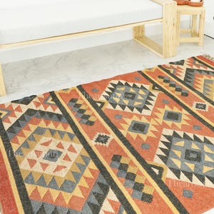 May include: A rectangular rug with a geometric pattern in red, grey, yellow, and black. The design features diamond and triangle shapes, giving a Southwestern or tribal feel. The rug is placed on a light-coloured floor, adding warmth to the room.