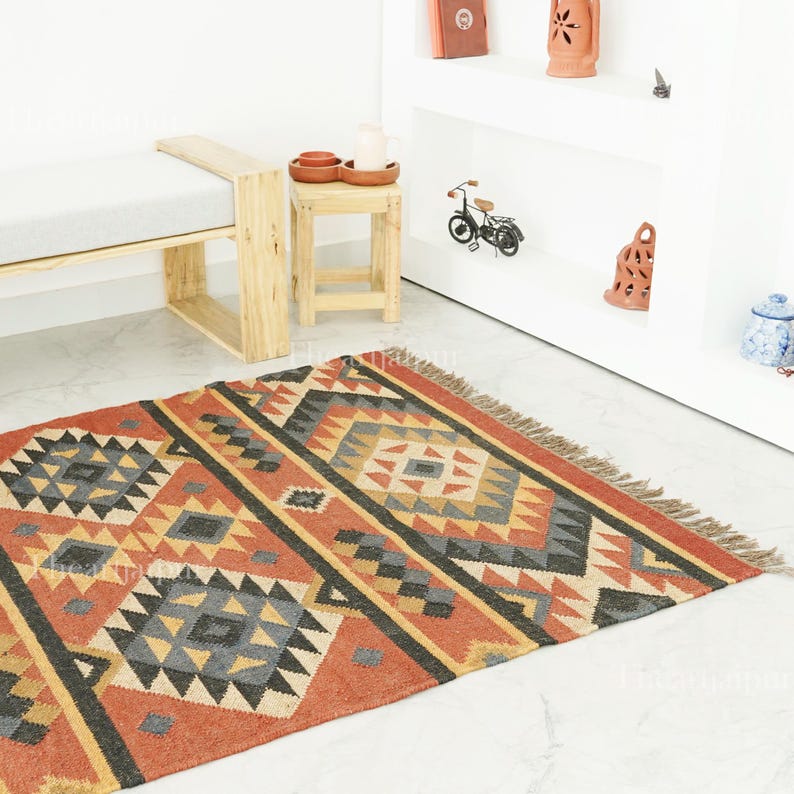 May include: A rectangular rug with a geometric pattern in red, orange, yellow, grey, and black. The rug has a fringed edge and is on a white tiled floor. The design features diamond and triangle shapes. The rug is suitable for a living room.