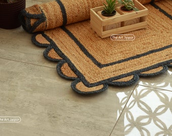 May include: A natural jute rug with a scalloped edge and a dark gray border. The rug is partially rolled up, revealing its texture and design. The Art Jaipur logo is visible.