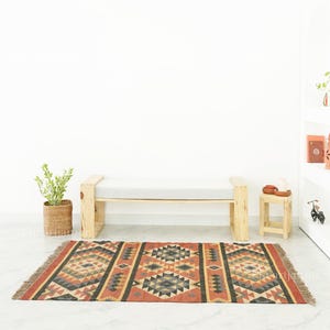 May include: A patterned rug with a geometric design in red, black, grey, and tan. It lies in front of a wooden bench with a grey cushion. A small wooden side table and a potted plant are also visible in the room.