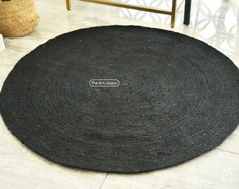 Puede incluir: Una alfombra redonda de yute negro con un patrón circular. La alfombra está hecha de fibras naturales y tiene un aspecto rústico.