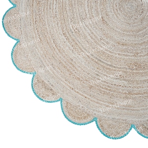Op de afbeelding: Een ronde jute vloerkleed met een geschulpte rand. Het vloerkleed is beige met een turquoise rand. De natuurlijke vezels creëren een textuur. Het ontwerp van het vloerkleed is zichtbaar in de hoek, met zijn ronde vorm en decoratieve rand.