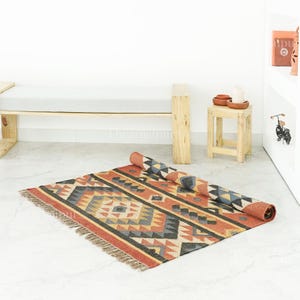 May include: A rolled-up, rectangular rug with a geometric pattern in shades of red, orange, grey, and beige. The rug has a fringed edge and is partially unrolled on a light-coloured floor. The rug is a decorative home decor item.