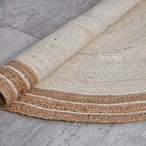 K&ouml;nnte beinhalten: Ein runder Naturfaser-Teppich mit einem wei&szlig;en Zentrum und einem braunen Jute-Rand. Der Teppich ist aufgerollt und zeigt die Textur der Jute.