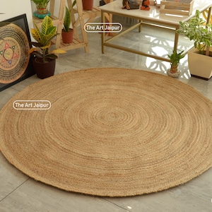 Könnte beinhalten: Ein großer, runder Jute-Teppich in natürlicher, hellbrauner Farbe. Der Teppich hat ein Spiralmuster und liegt auf einem gefliesten Boden. The Art Jaipur ist auf dem Teppich aufgedruckt.