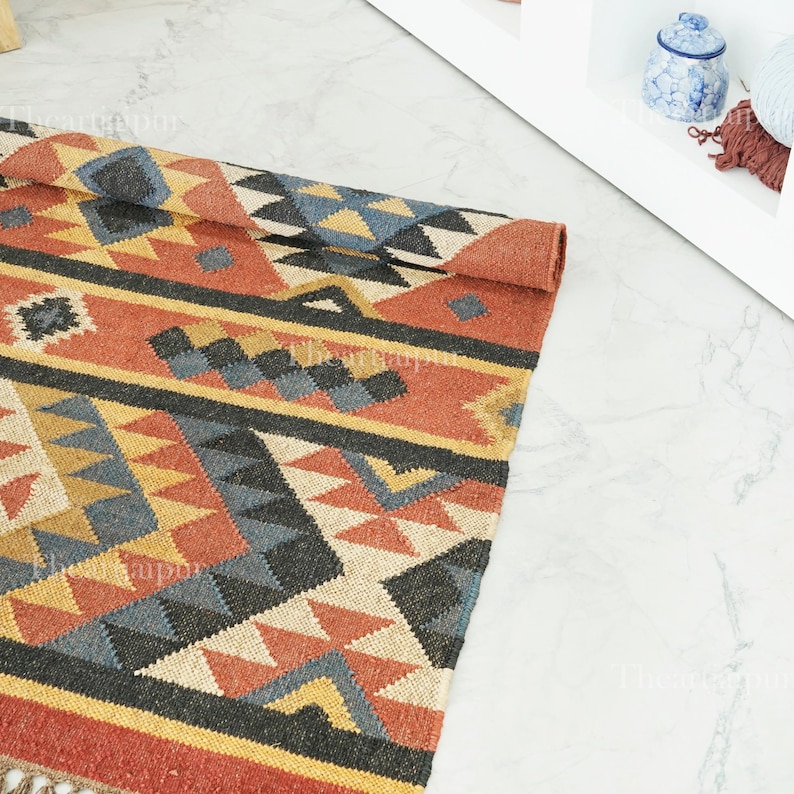 May include: A rolled-up, rectangular rug with a geometric pattern in shades of red, yellow, black, and blue. The rug has a fringe along one edge and is made of a woven material. The design features triangles and diamond shapes.