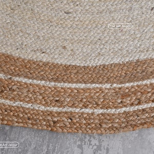 K&ouml;nnte beinhalten: Ein runder Jute-Teppich mit einem wei&szlig;en Zentrum und einem braunen Rand mit wei&szlig;en Streifen. Der Teppich ist aus Naturfasern gefertigt und hat einen rustikalen Look.