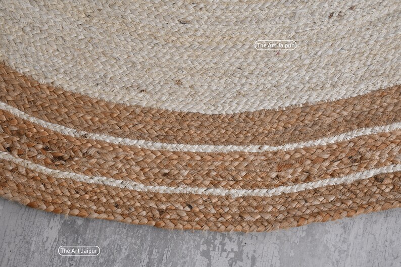 K&ouml;nnte beinhalten: Ein runder Jute-Teppich mit einem wei&szlig;en Zentrum und einem braunen Rand mit wei&szlig;en Streifen. Der Teppich ist aus Naturfasern gefertigt und hat einen rustikalen Look.
