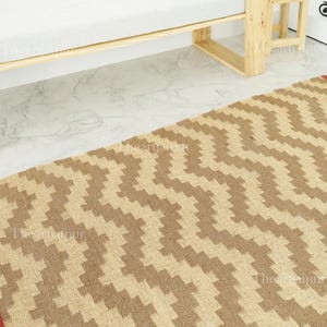 Puede incluir: Una alfombra tejida con un patrón en zigzag en tonos marrón y beige. La alfombra está colocada sobre un suelo de mármol, con un marco de cama de madera visible en el fondo. El diseño de la alfombra añade textura y calidez al espacio.