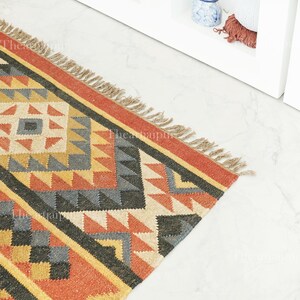 May include: A colourful geometric rug featuring a repeating pattern of triangles and squares in shades of red, orange, yellow, grey, and black. The rug has a fringed edge and is made from a woven material. The rug is suitable for a hallway or living room.