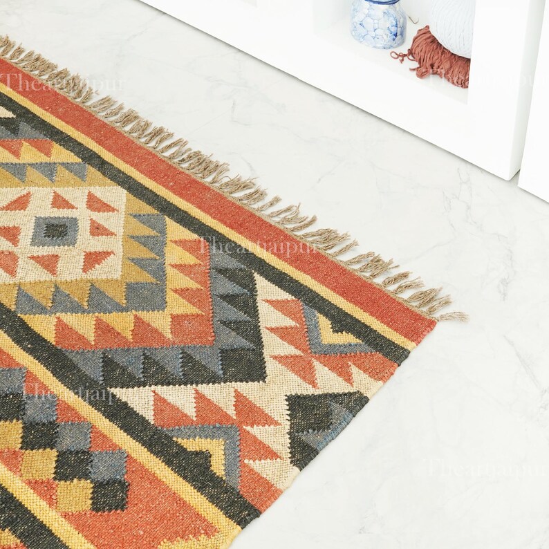 May include: A colourful geometric rug featuring a repeating pattern of triangles and squares in shades of red, orange, yellow, grey, and black. The rug has a fringed edge and is made from a woven material. The rug is suitable for a hallway or living room.