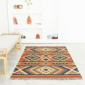 May include: A rectangular rug with a geometric pattern in red, orange, black, grey, and yellow. The rug has a fringed edge and is placed on a white floor. The design features diamond and triangle shapes. The rug measures approximately 180 cm x 120 cm.