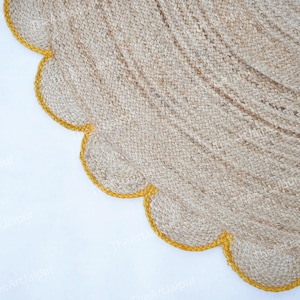 Op de afbeelding: Een ronde jute vloerkleed met een geschulpte rand. Het vloerkleed heeft een natuurlijke beige kleur met een heldergele rand. De textuur van het vloerkleed is zichtbaar, met de geweven vezels. Het vloerkleed ligt op een witte achtergrond.
