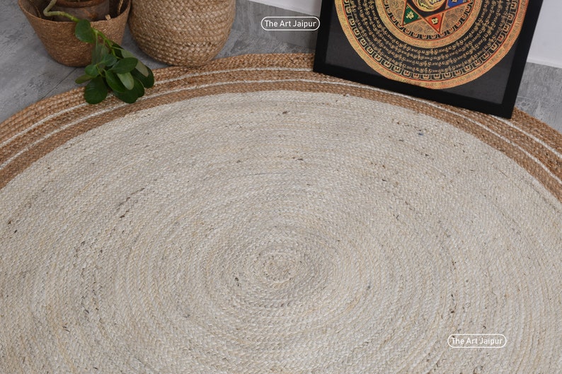 K&ouml;nnte beinhalten: Ein runder Jute-Teppich mit einem wei&szlig;en Zentrum und einem braunen Rand. Der Teppich hat eine nat&uuml;rliche, gewebte Textur. Der Text "The Art Jaipur" ist in der unteren rechten Ecke des Bildes sichtbar.