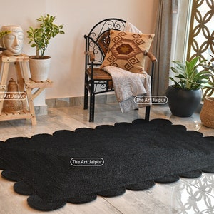 May include: Black jute rug with a scalloped edge. The rug is on a light wood floor in a room with a chair, plants, and a window.