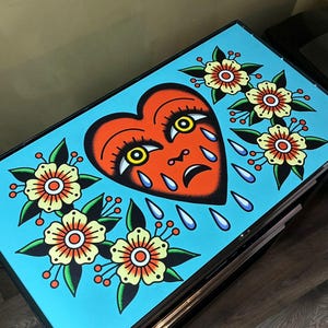 May include: A rectangular metal storage cabinet featuring a vibrant light blue top surface decorated with a traditional tattoo-style illustration of a crying red heart with yellow eyes, surrounded by clusters of yellow and orange flowers.
