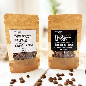 Op de afbeelding: Twee bruine papieren zakjes met witte en zwarte etiketten. De etiketten lezen "The Perfect Blend" met de namen "Sarah & Tom" en de datum "09.10.23". De zakjes zijn gevuld met koffiebonen en staan op een wit houten oppervlak. Er liggen koffiebonen verspreid rond de zakjes.