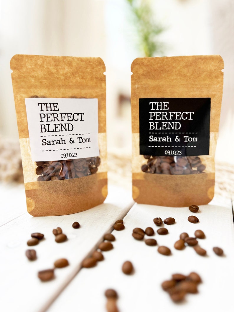 Op de afbeelding: Twee bruine papieren zakjes met witte en zwarte etiketten. De etiketten lezen "The Perfect Blend" met de namen "Sarah & Tom" en de datum "09.10.23". De zakjes zijn gevuld met koffiebonen en staan op een wit houten oppervlak. Er liggen koffiebonen verspreid rond de zakjes.