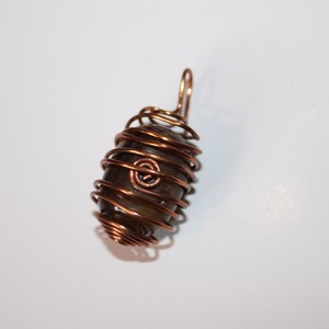 May include: A dark brown stone wrapped in copper wire, forming a pendant. The wire is twisted and coiled around the stone, creating a cage-like effect.