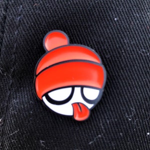 May include: A red and white enamel pin of a cartoon character wearing a red beanie hat with a white face and a red tongue sticking out.
