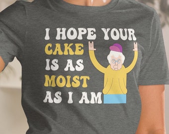 Peut inclure: T-shirt gris foncé avec le texte "I HOPE YOUR CAKE IS AS MOIST AS I AM" et une image de dessin animé d'une femme âgée les bras levés. Le t-shirt est fait d'une matière douce.