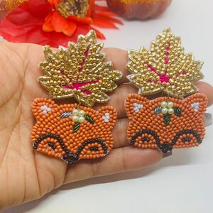 K&ouml;nnte beinhalten: Ein Paar Perlenohrringe mit einem Fuchs- und Blattdesign. Der Fuchs ist orange mit schwarzen und wei&szlig;en Details. Das Blatt ist goldfarben mit rosa Akzenten. Die Ohrringe sind handgefertigt und f&uuml;r den Herbst geeignet.