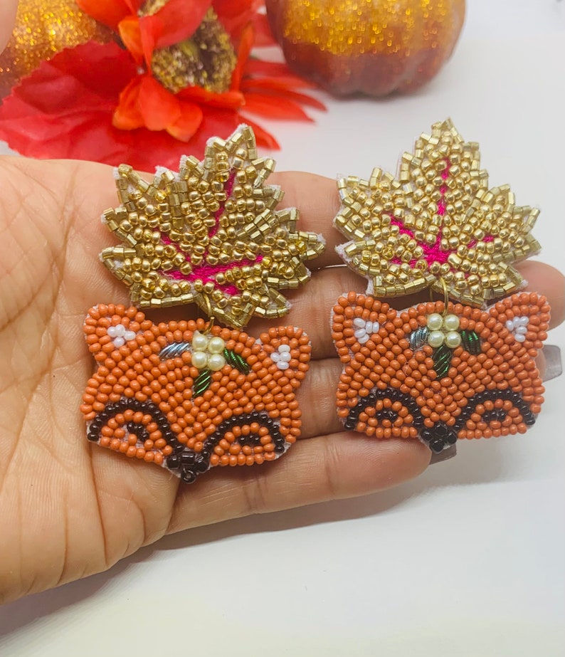 K&ouml;nnte beinhalten: Ein Paar Perlenohrringe mit einem Fuchs- und Blattdesign. Der Fuchs ist orange mit schwarzen und wei&szlig;en Details. Das Blatt ist goldfarben mit rosa Akzenten. Die Ohrringe sind handgefertigt und f&uuml;r den Herbst geeignet.