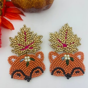 K&ouml;nnte beinhalten: Ein Paar bestickte Fuchsohrringe. Die Ohrringe zeigen ein Fuchgesicht in Orange- und Schwarzt&ouml;nen, gekr&ouml;nt von einem goldfarbenen Blatt. Die Bl&auml;tter haben ein rosa Aderdesign. Kleine wei&szlig;e Blumen befinden sich auf dem Kopf des Fuchses.