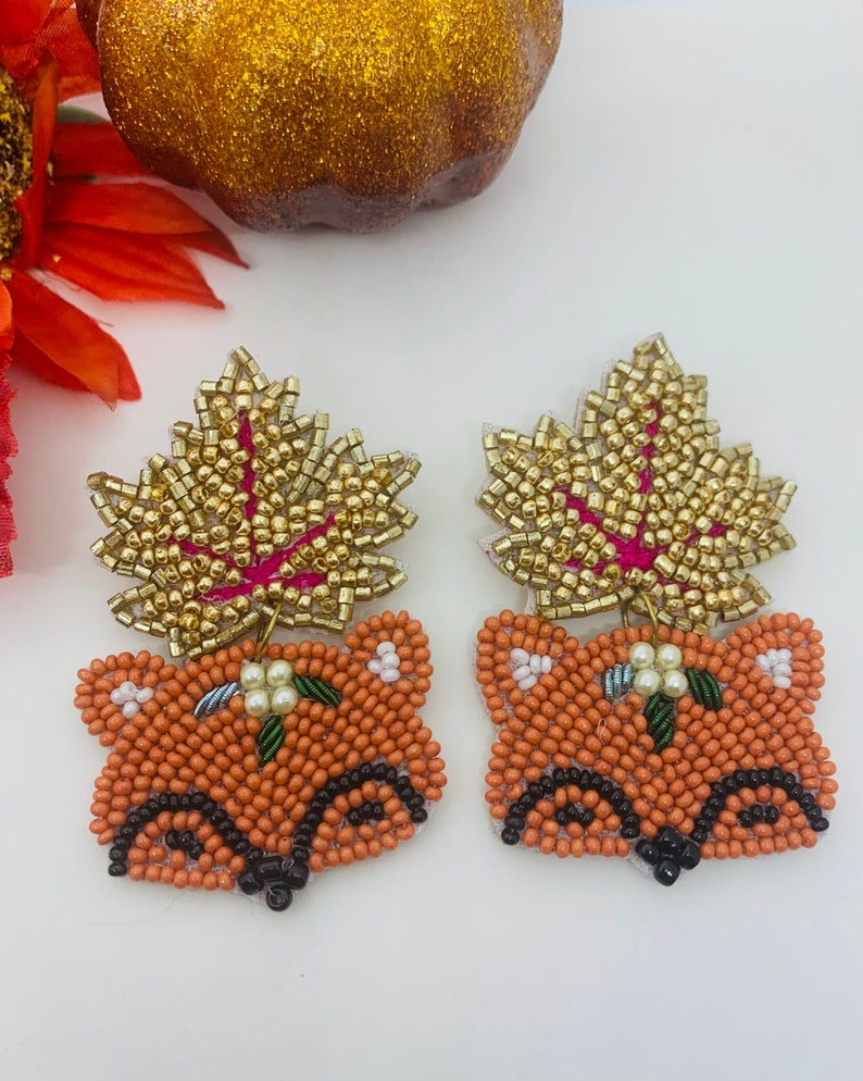 K&ouml;nnte beinhalten: Ein Paar bestickte Fuchsohrringe. Die Ohrringe zeigen ein Fuchgesicht in Orange- und Schwarzt&ouml;nen, gekr&ouml;nt von einem goldfarbenen Blatt. Die Bl&auml;tter haben ein rosa Aderdesign. Kleine wei&szlig;e Blumen befinden sich auf dem Kopf des Fuchses.