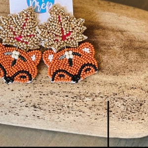K&ouml;nnte beinhalten: Perlenohrringe mit einem Fuchskopf-Design in Orange, Schwarz und Wei&szlig;. Die Ohrringe haben ein blattf&ouml;rmiges Oberteil in Gold und Rot. Die Ohrringe werden auf einer Holzoberfl&auml;che pr&auml;sentiert.