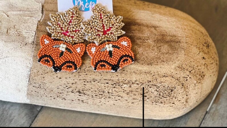 K&ouml;nnte beinhalten: Perlenohrringe mit einem Fuchskopf-Design in Orange, Schwarz und Wei&szlig;. Die Ohrringe haben ein blattf&ouml;rmiges Oberteil in Gold und Rot. Die Ohrringe werden auf einer Holzoberfl&auml;che pr&auml;sentiert.