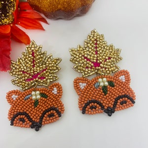 K&ouml;nnte beinhalten: Ein Paar bestickte Fuchsohrringe mit goldenen Bl&auml;ttern. Die Ohrringe zeigen orangefarbene Fuchsk&ouml;pfe mit schwarzen Details und goldperlenbesetzten Bl&auml;ttern mit rosa R&uuml;ckseite. Kleine wei&szlig;e Blumen und gr&uuml;ne Bl&auml;tter sind ebenfalls vorhanden.