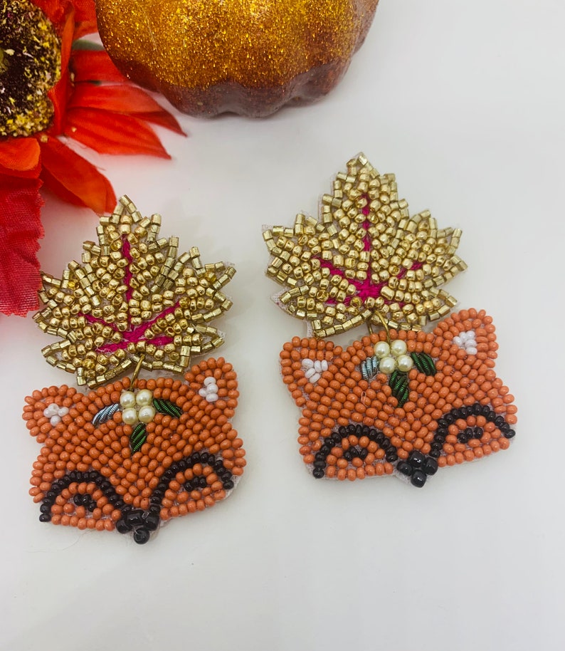 K&ouml;nnte beinhalten: Ein Paar bestickte Fuchsohrringe mit goldenen Bl&auml;ttern. Die Ohrringe zeigen orangefarbene Fuchsk&ouml;pfe mit schwarzen Details und goldperlenbesetzten Bl&auml;ttern mit rosa R&uuml;ckseite. Kleine wei&szlig;e Blumen und gr&uuml;ne Bl&auml;tter sind ebenfalls vorhanden.