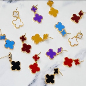 May include: Assortment of colourful clover-shaped earrings with gold accents. The earrings are in various colours, including red, blue, purple, black, white, and yellow.