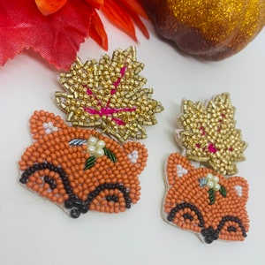 K&ouml;nnte beinhalten: Ein Paar bestickte Fuchsohrringe mit goldenen Blattakzenten. Die Fuchsk&ouml;pfe sind orange mit schwarzen Details und wei&szlig;en Akzenten. Die Bl&auml;tter sind goldfarben mit rosa Adern. Die Ohrringe liegen auf einer wei&szlig;en Oberfl&auml;che.