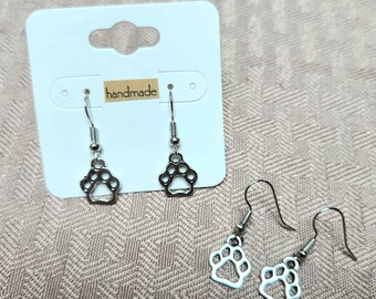 May include: Silver-colored paw print earrings with a fish hook design. The earrings are displayed on a white card with a small brown label that reads "handmade". The earrings are a pet-themed accessory.