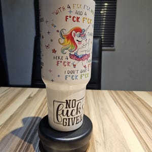 Op de afbeelding: Een witte roestvrijstalen beker met een doorzichtig deksel, voorzien van een kleurrijke eenhoorngrafiek in een roze tutu die de middelvinger opsteekt. De tekst luidt "WITH A F*CK F*CK HERE AND A F*CK F*CK THERE" en "NO F*CKS GIVEN" met sterretjes.