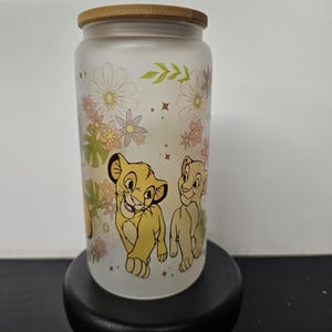 Könnte beinhalten: Ein Glasbecher mit Bambusdeckel, mit Simba und Nala aus Der König der Löwen. Der Becher ist mit floralen Akzenten in Rosa, Gelb und Grün verziert. Der Sockel ist schwarz.