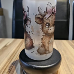 Könnte beinhalten: Ein mattiertes Glas mit Bambusdeckel und einer Cartoon-Illustration eines flauschigen braunen Hochlandrindes mit rosa Schleife, das aus einem Becher mit Strohhalm trinkt. Das Design zeigt braunes Fell, große Augen und kleine Hörner.