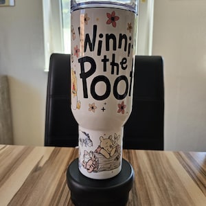 Può includere: Un bicchiere cilindrico bianco con coperchio trasparente che riporta la scritta nera Winnie the Pooh, circondata da piccole illustrazioni di fiori arancioni e rosa. La parte inferiore mostra un disegno di Winnie the Pooh, Pimpi e Ih-Oh su un tronco.