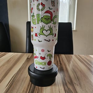 Könnte beinhalten: Ein weißer Edelstahlbecher mit einem wiederkehrenden Weihnachtsmuster aus grünen Grinch-Figuren, roten Weihnachtsmützen, Zuckerstangen, Kränzen und Geschenkkartons. Der Schriftzug "GRINCH" ist in Grün gedruckt und der Becher hat einen Kunststoffdeckel.